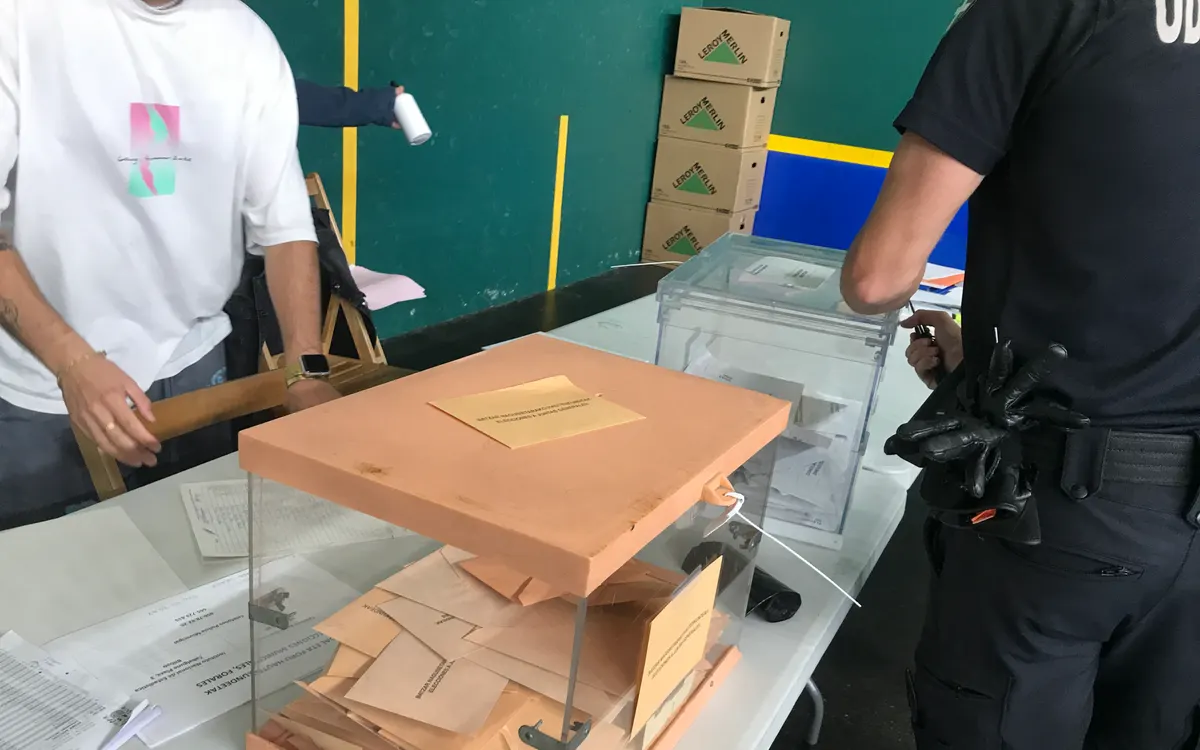 Urna transparente de tapa naranja con sobres de votación sobre una mesa en un colegio electoral