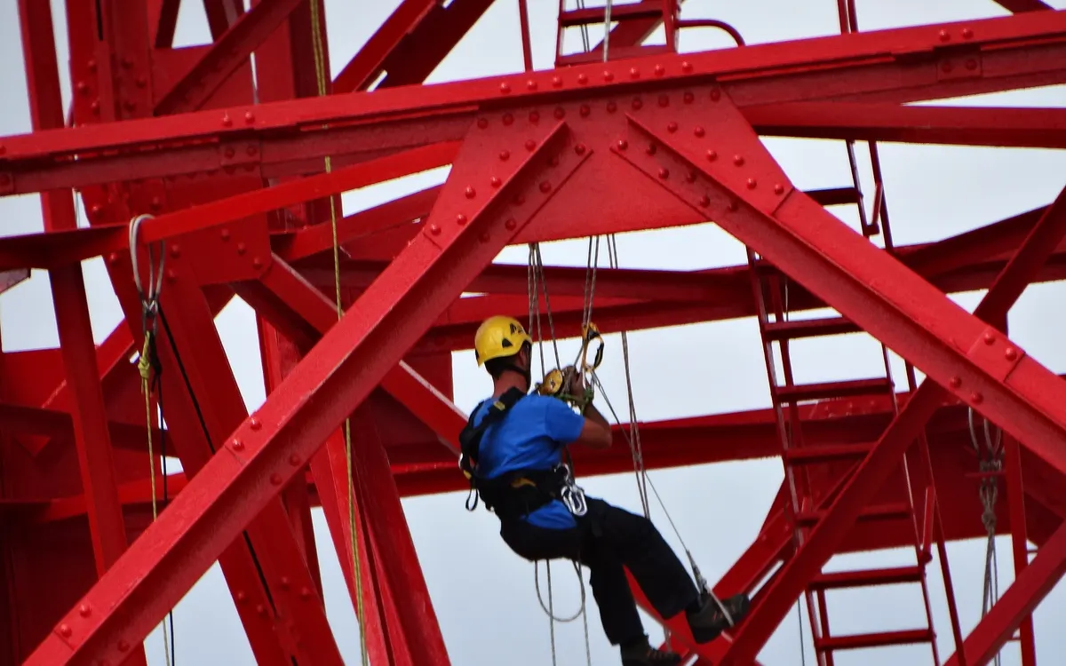 Trabajador con casco colgado de cuerdas entre vigas rojas de acero en una estructura